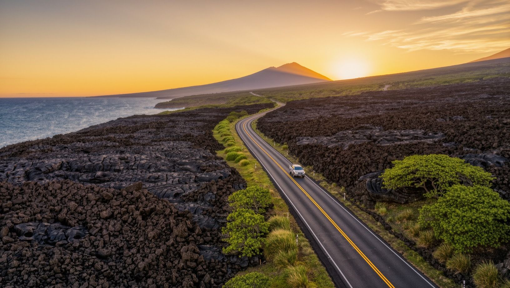 A highway winding between black lava fields and the Pacific at sunset under Mauna Kea, a pickup truck in the distance